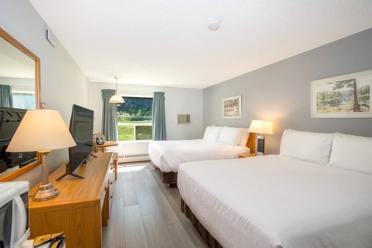 A hotel room with two beds, a desk with a lamp, a TV, and a large window with curtains.