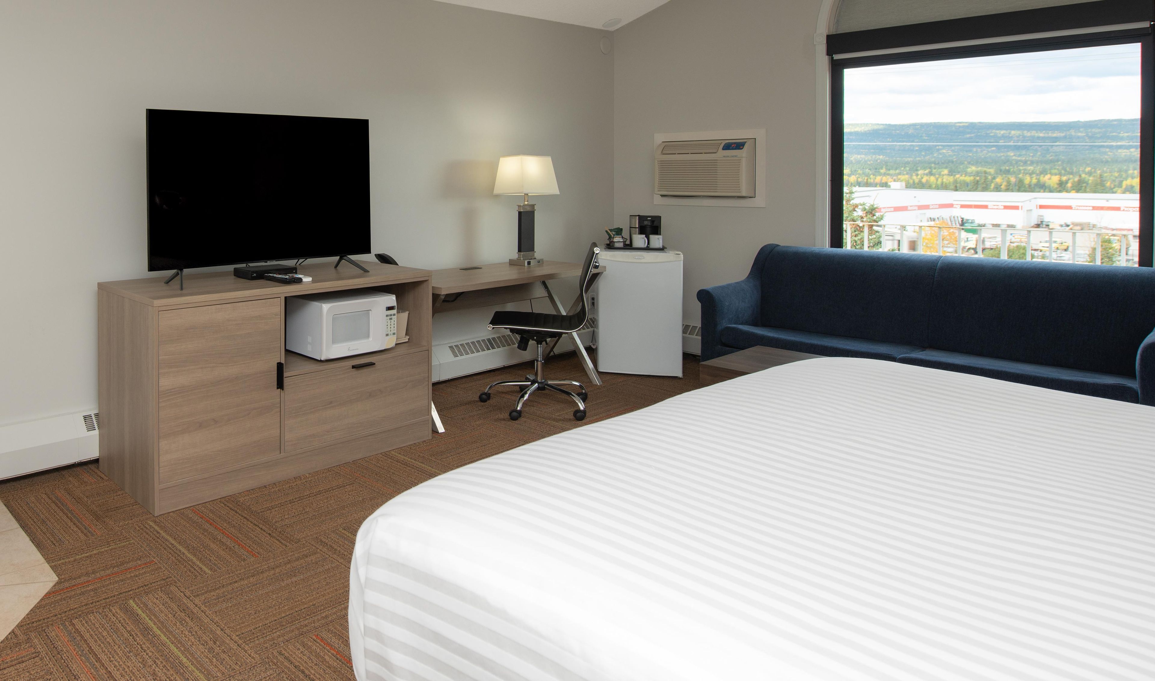 A hotel room with a bed, a desk with a computer, a TV, a lamp, a coffee machine, and a view of the city.