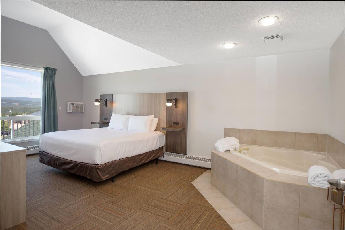 A modern hotel room with a large bed, a separate bathroom with a bathtub, and a view of the outdoors.