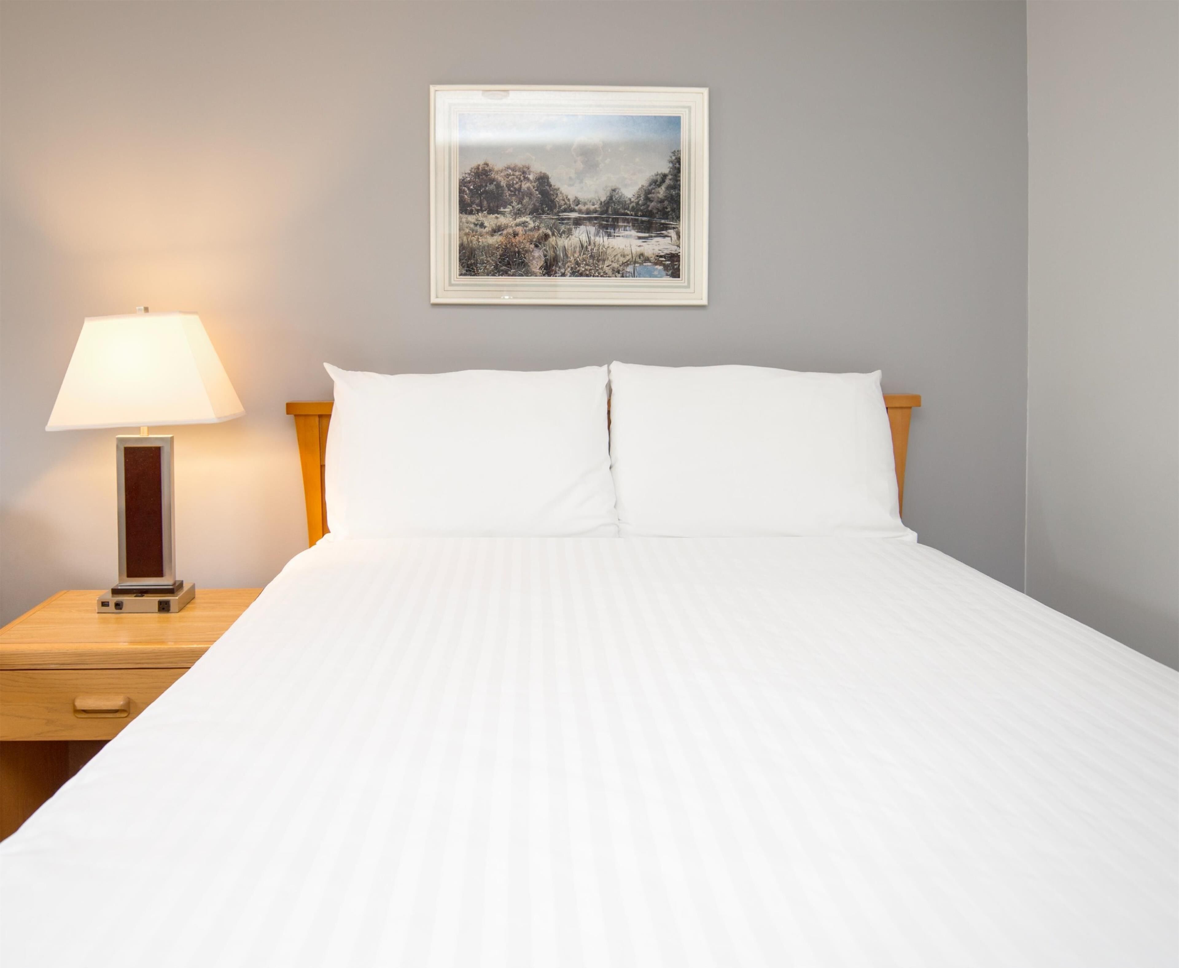 A neatly made bed with white linens, a framed picture of a landscape on the wall, and a bedside table with a lamp.