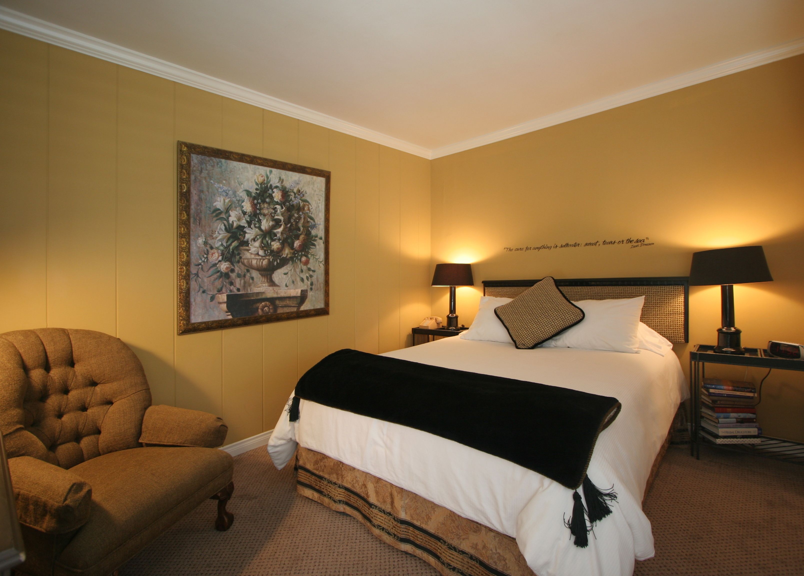 A hotel room with a large bed, two bedside lamps, a chair, a painting on the wall, and a bookshelf with books.