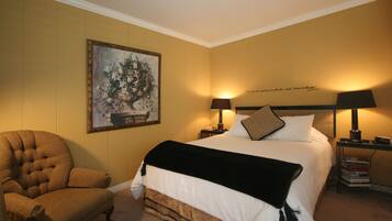 A hotel room with a large bed, two bedside lamps, a chair, a painting on the wall, and a bookshelf with books.
