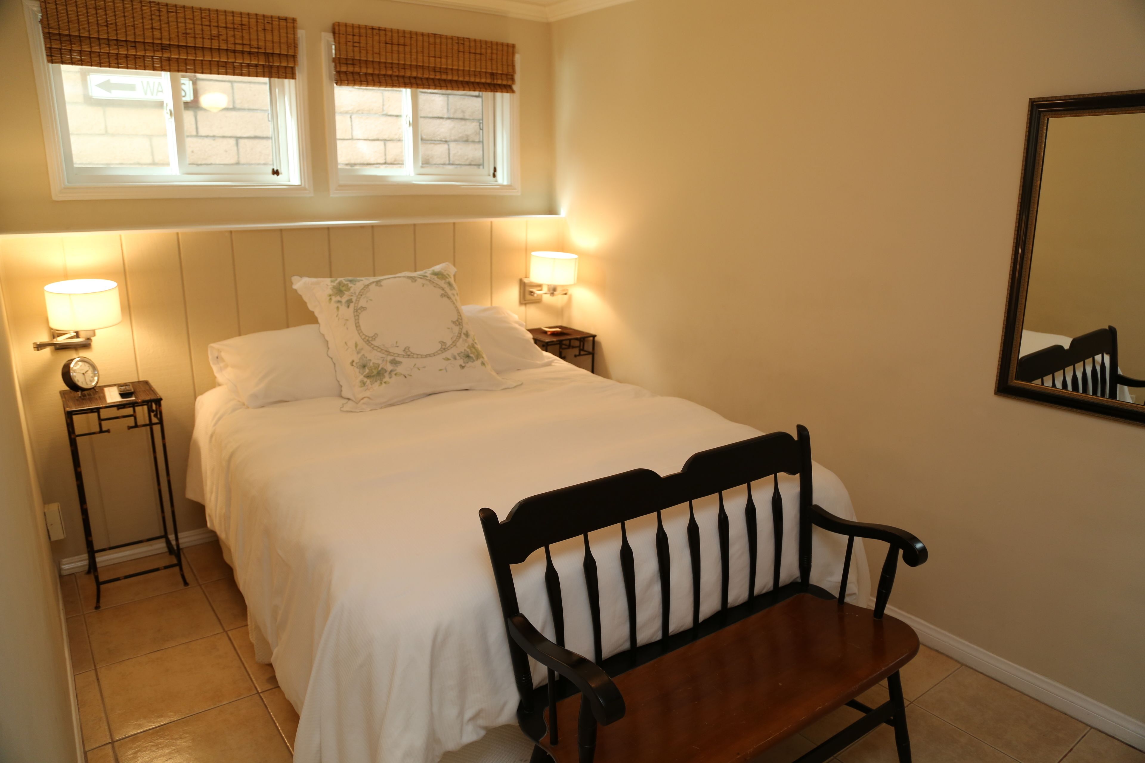 A neatly made bed with white bedding, two bedside lamps, a wooden chair, and a wall-mounted mirror.