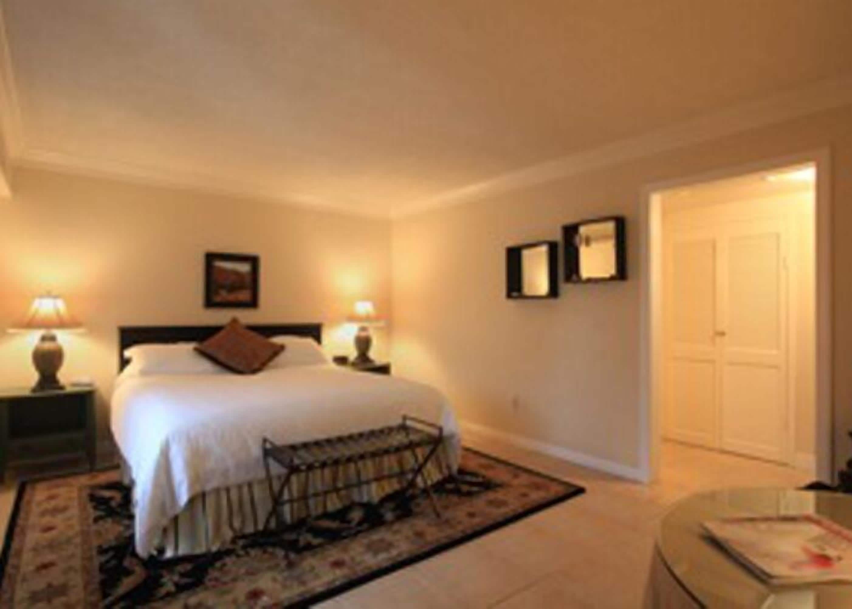 A hotel room with a bed, bedside lamps, a small table, a chair, and a door.