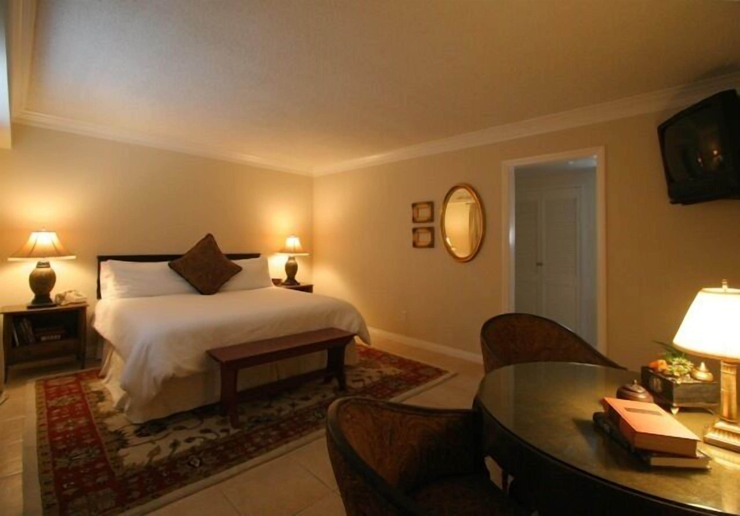 A hotel room with a bed, bedside tables with lamps, a chair, a small table with a lamp, a mirror, and a television on the wall.