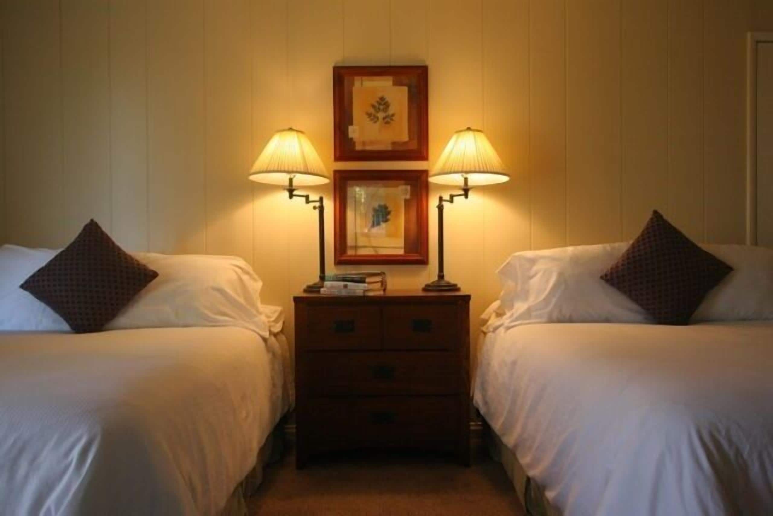 Une chambre d’hôtel avec deux lits, chacun muni d’un oreiller et d’une table de chevet avec une lampe. Un tableau encadré est accroché au mur au-dessus des tables de chevet.