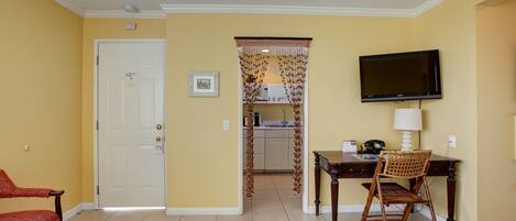 A hotel room with a bed, a desk with a chair, a television, and a view into the kitchen.