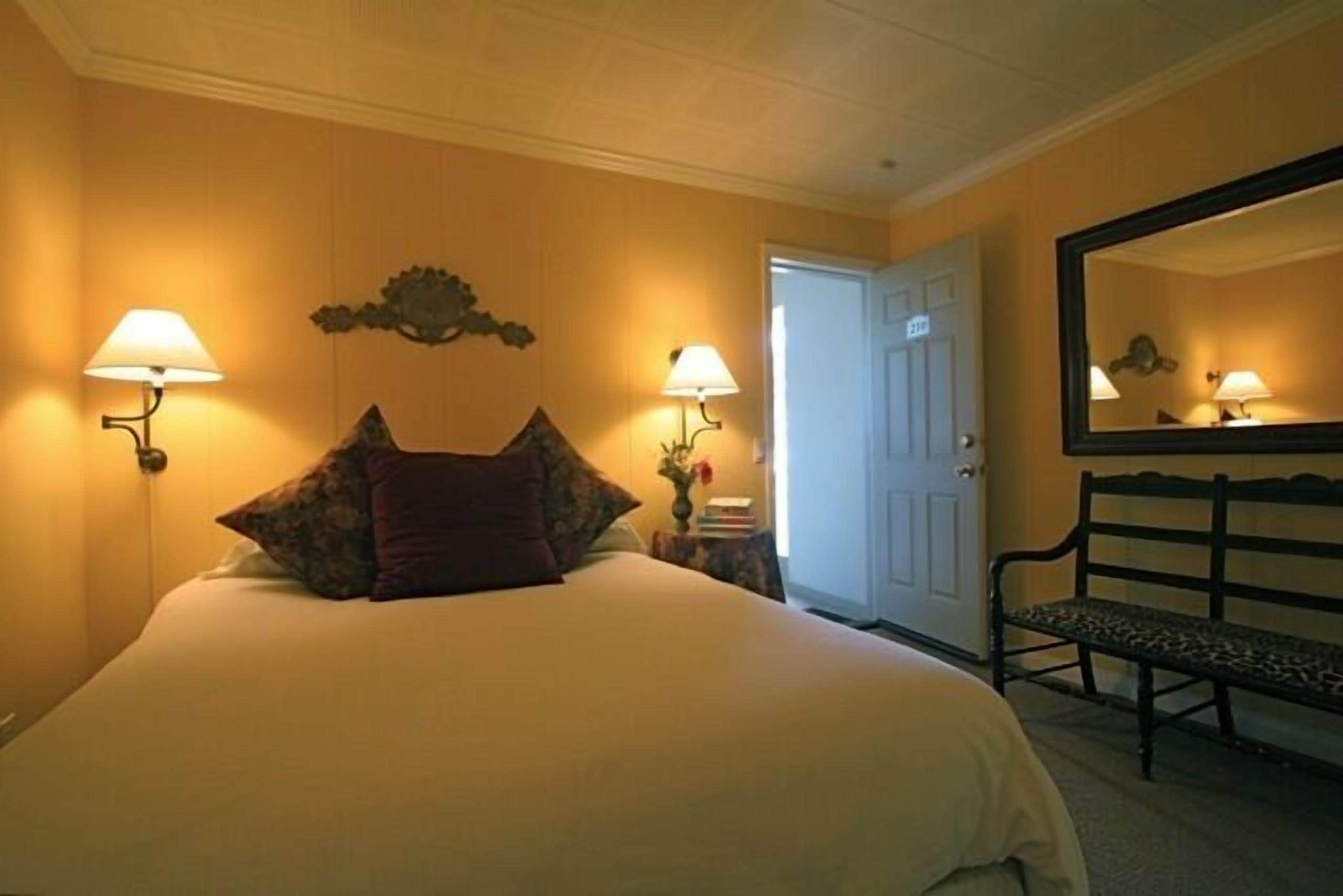 A hotel room with a large bed, two bedside lamps, a bench, and a door.