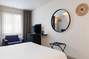 Habitación de hotel con cama, televisión, sillón, escritorio con planta, espejo y estante de pared con fotografías.