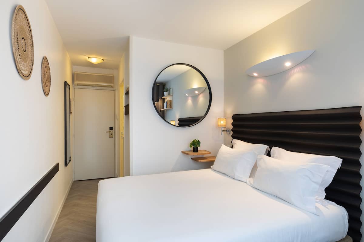 Una habitación de hotel con una cama, un espejo redondo, una mesita de noche con una planta y un aplique de pared.