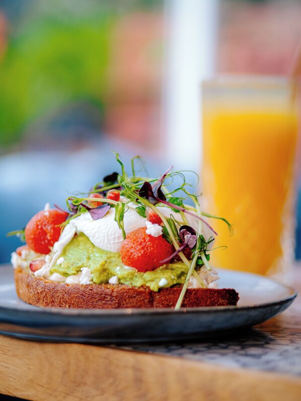 Un sándwich de aguacate, tomates y verduras sobre una rebanada de pan, acompañado de un vaso de jugo de naranja.