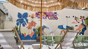 Un espacio de descanso frente a la playa, con sillones a rayas, un mural con palmeras y una escena costera, y una sombrilla de paja.