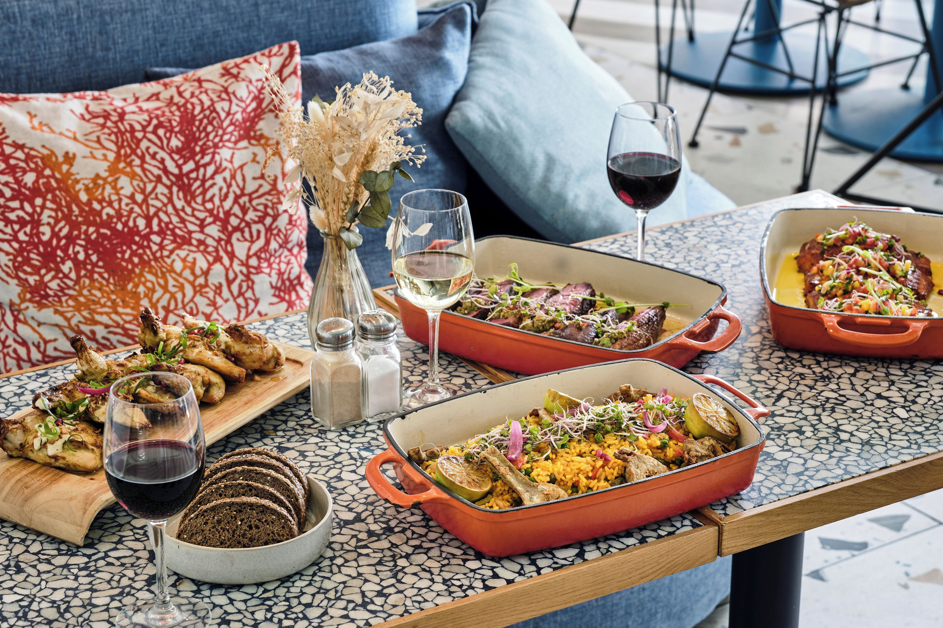 A dining table with two red enameled cast iron dishes containing food, a glass of red wine, and a bowl of bread.