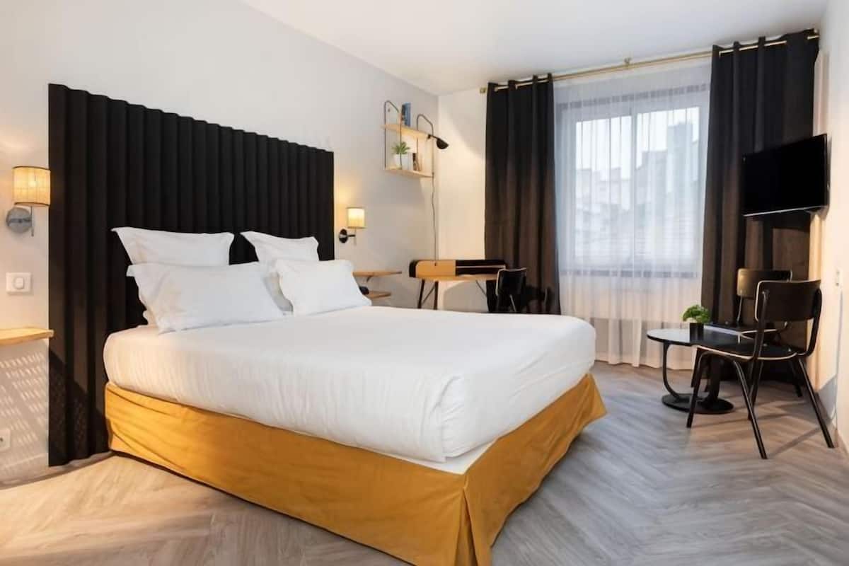 Habitación de hotel con cama, escritorio, silla, televisor y ventana con cortinas.