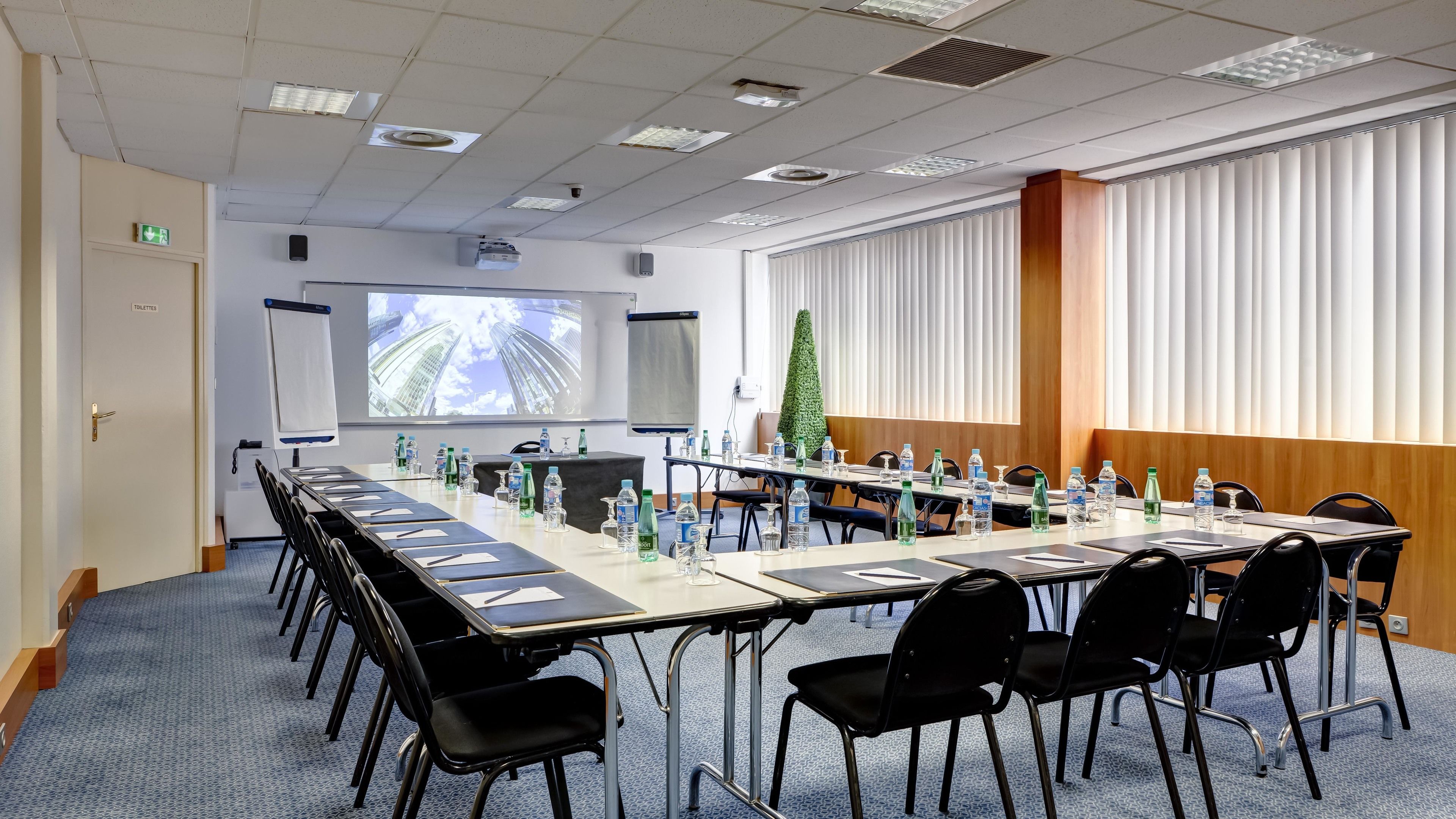 Un salón de conferencias con una larga mesa, sillas, una pantalla de proyector, un dispensador de agua y una pequeña planta.