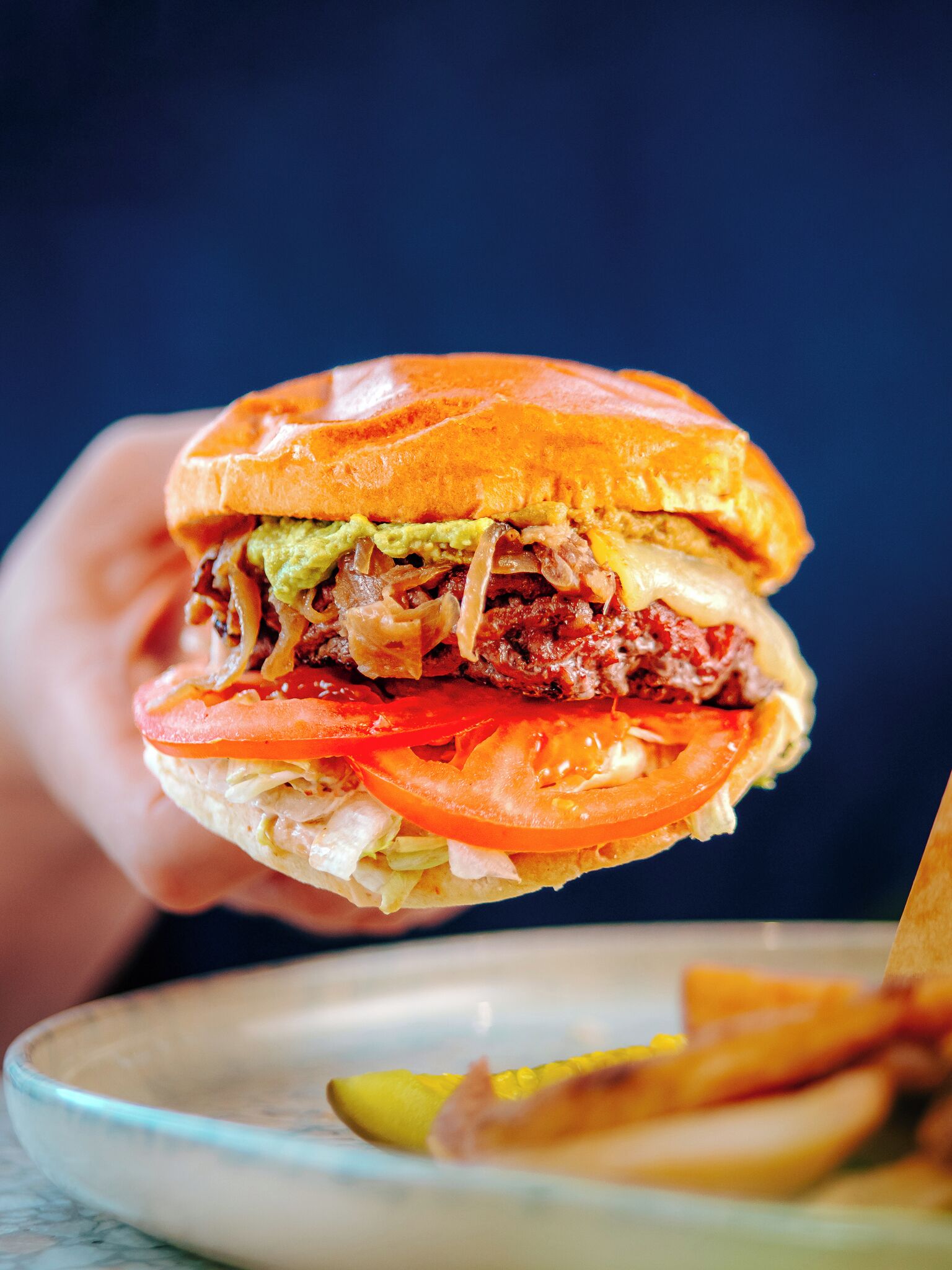 Un gros plan d’une main tenant un hamburger, où l’on peut voir clairement les ingrédients, comme la laitue, la tomate et le fromage.
