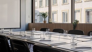 Un salón de conferencias con una larga mesa, sillas, una pantalla de proyector y vistas a un edificio con ventanas.