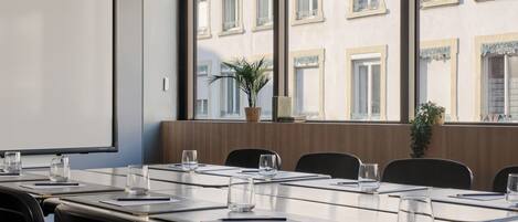 Een vergaderruimte met een lange tafel, stoelen, een projectorscherm en uitzicht op een gebouw met ramen.