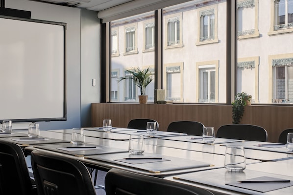 Un salón de conferencias con una larga mesa, sillas, una pantalla de proyector y vistas a un edificio con ventanas.