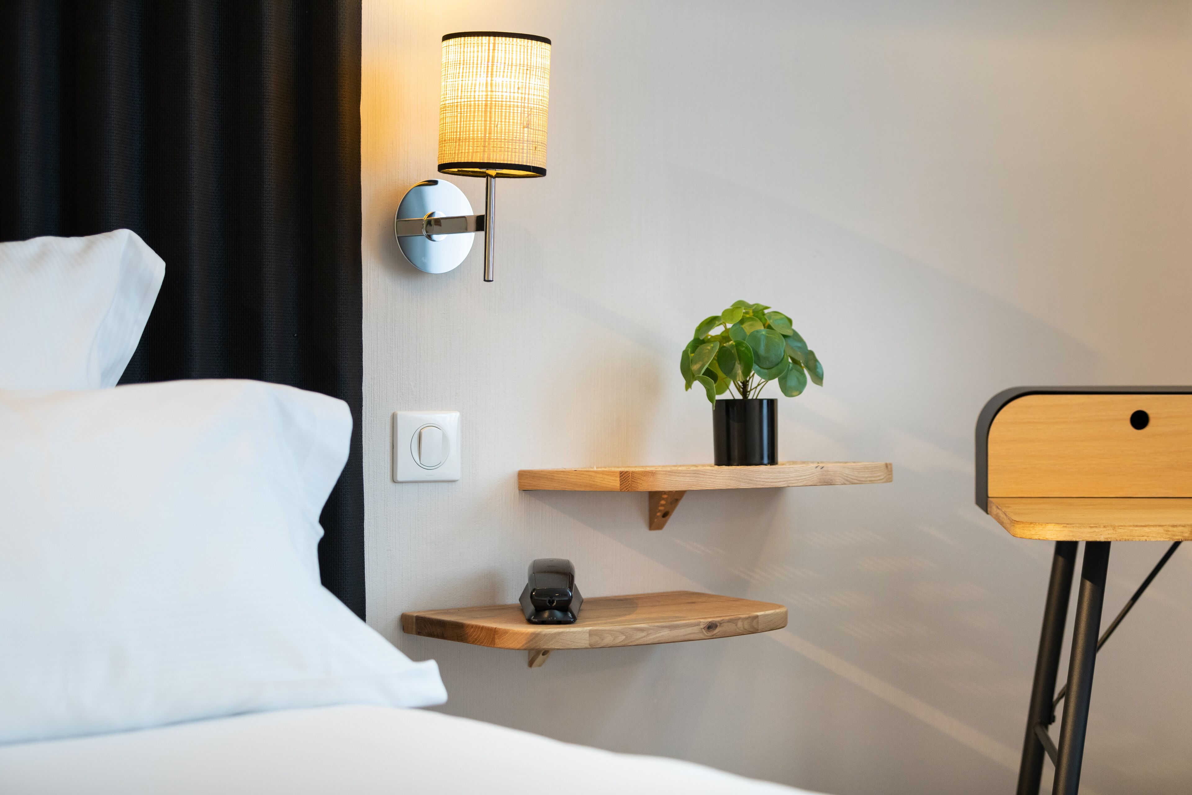 Una habitación de hotel con cama, lámpara de pared, estantes de madera y una pequeña planta.