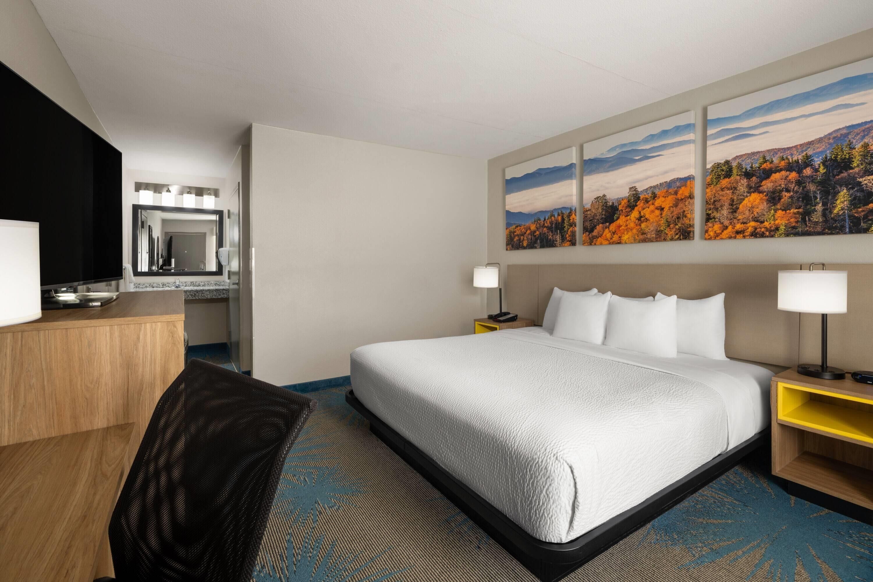A hotel room with a large bed, a desk with a mirror, and a painting of autumn foliage on the wall.