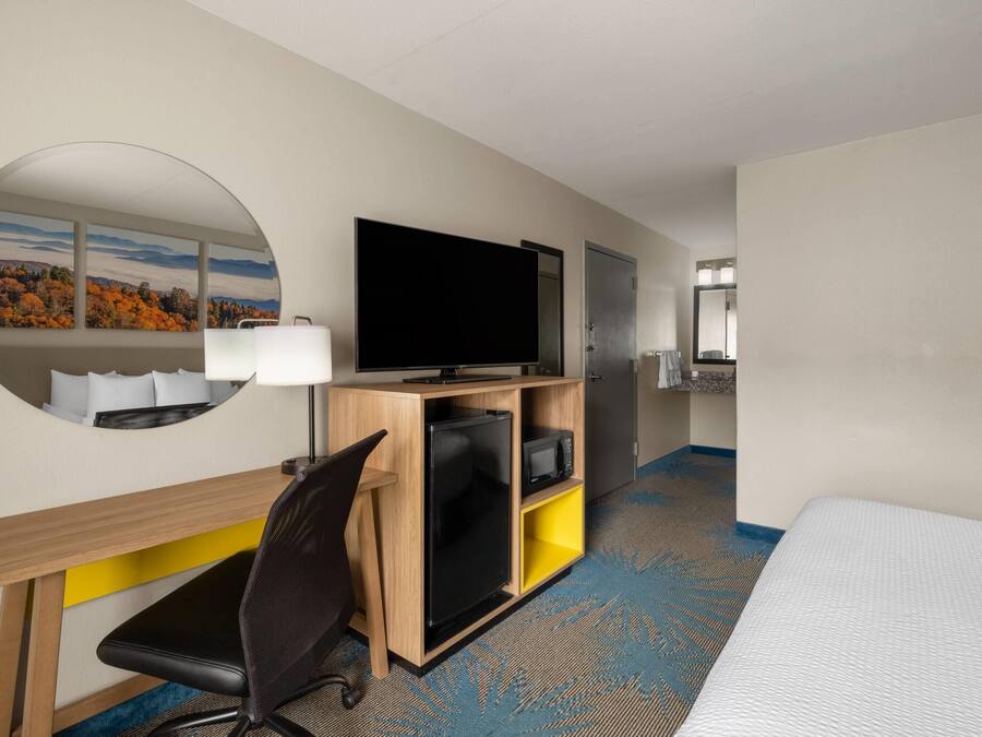 A hotel room with a bed, a desk with a flat-screen TV, a mirror, and a view of autumn trees.