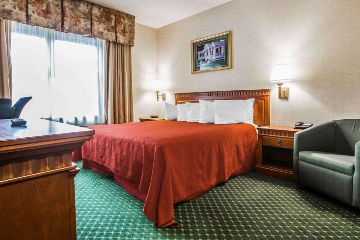 A hotel room with a bed, bedside tables, a desk, a chair, a window with curtains, and a wall-mounted picture.