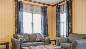 A living room with a sofa, armchair, and coffee table, two windows with curtains, and a tiled floor.