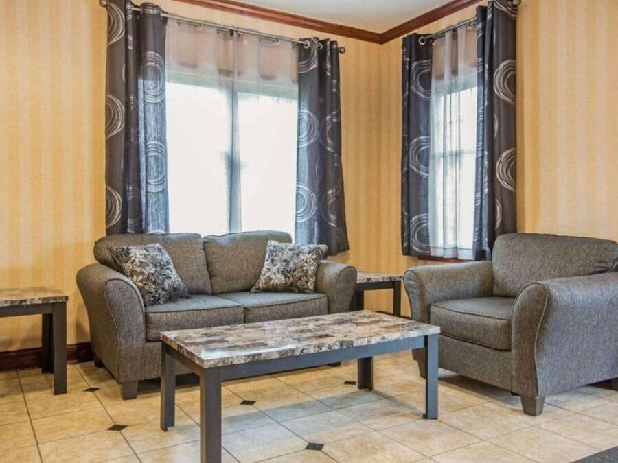 Ein Wohnzimmer mit Sofa, Sessel und Couchtisch, zwei Fenstern mit Vorhängen und einem Fliesenboden.