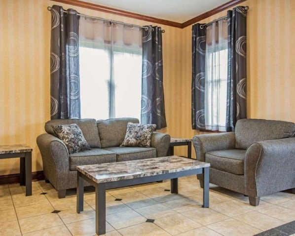 A living room with a sofa, armchair, and coffee table, two windows with curtains, and a tiled floor. - Point Hotel Queensbury (Queensbury)