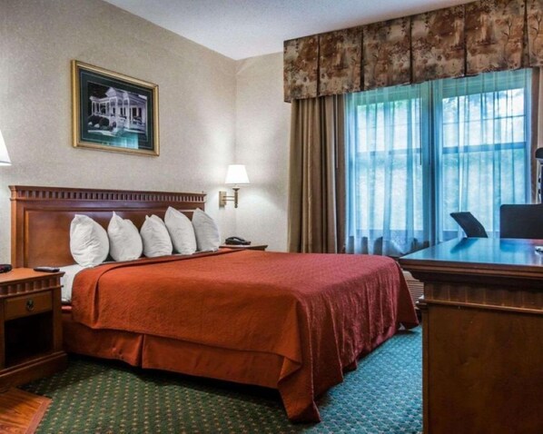 Habitación de hotel con una cama grande, un escritorio, una silla, una lámpara, un cuadro en la pared y una ventana con cortinas.