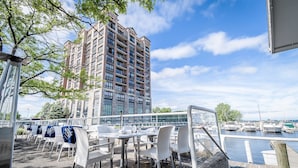An outdoor dining area with white chairs and tables, overlooking a marina with boats and a tall building in the background.
