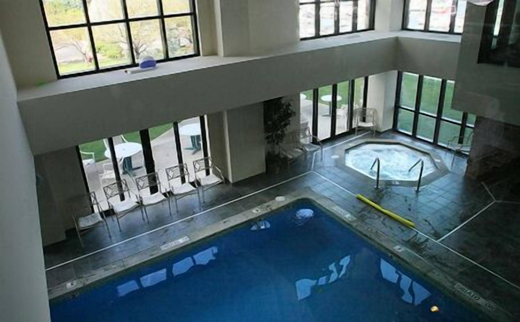 An indoor swimming pool with a hot tub, surrounded by large windows and outdoor seating.