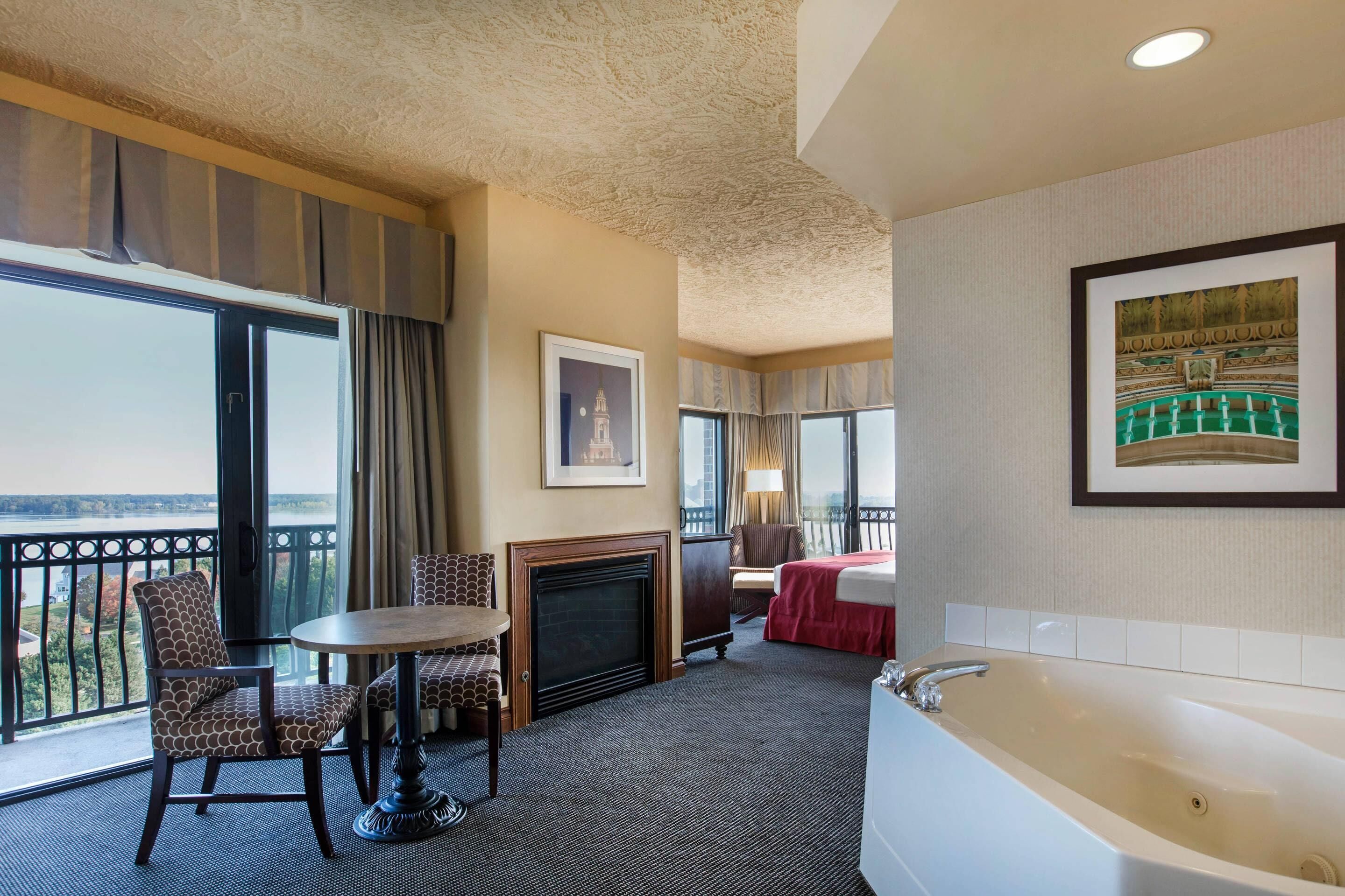A hotel room with a balcony, a fireplace, a large bathtub, and a dining area with a table and chairs.