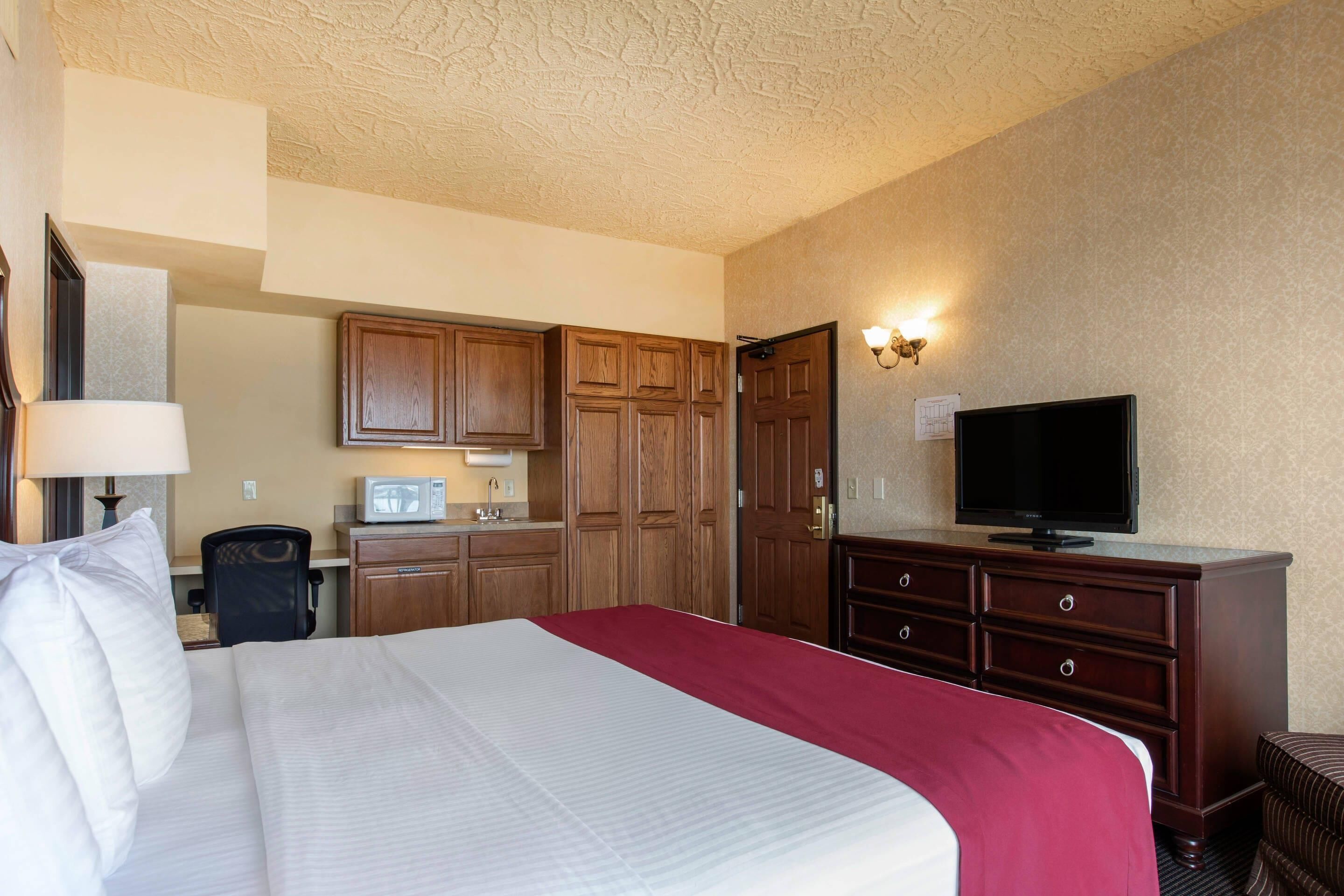 Quarto de hotel com uma cama grande, televisão, armários de madeira e uma kitchenette.