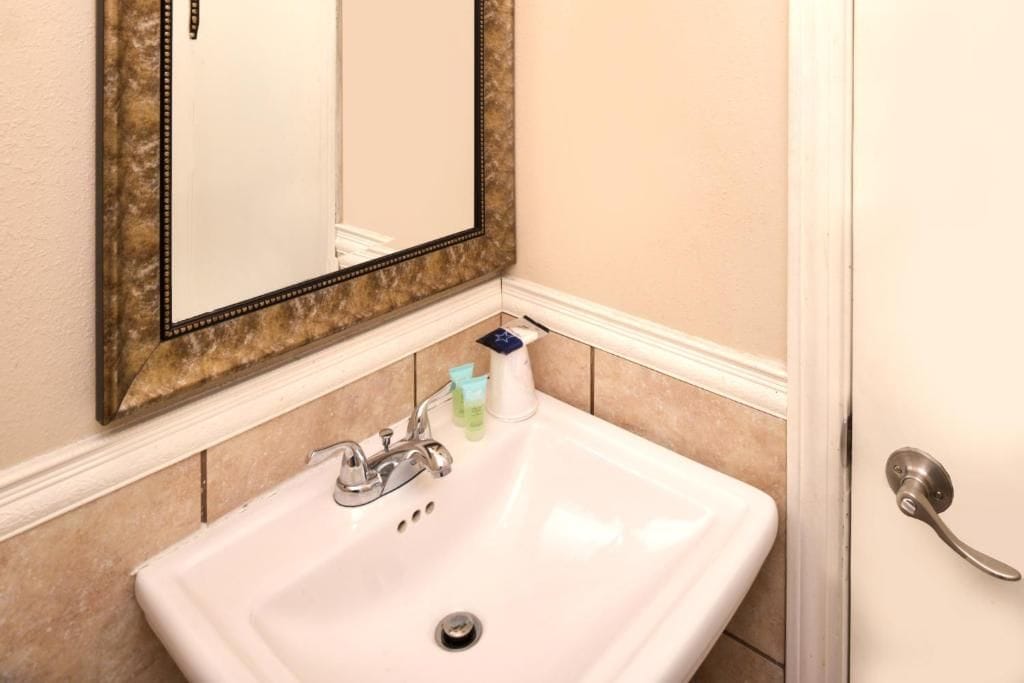 Un bany amb un rentapans blanc, un mirall amb un marc decoratiu i una porta amb una nansa platejada.