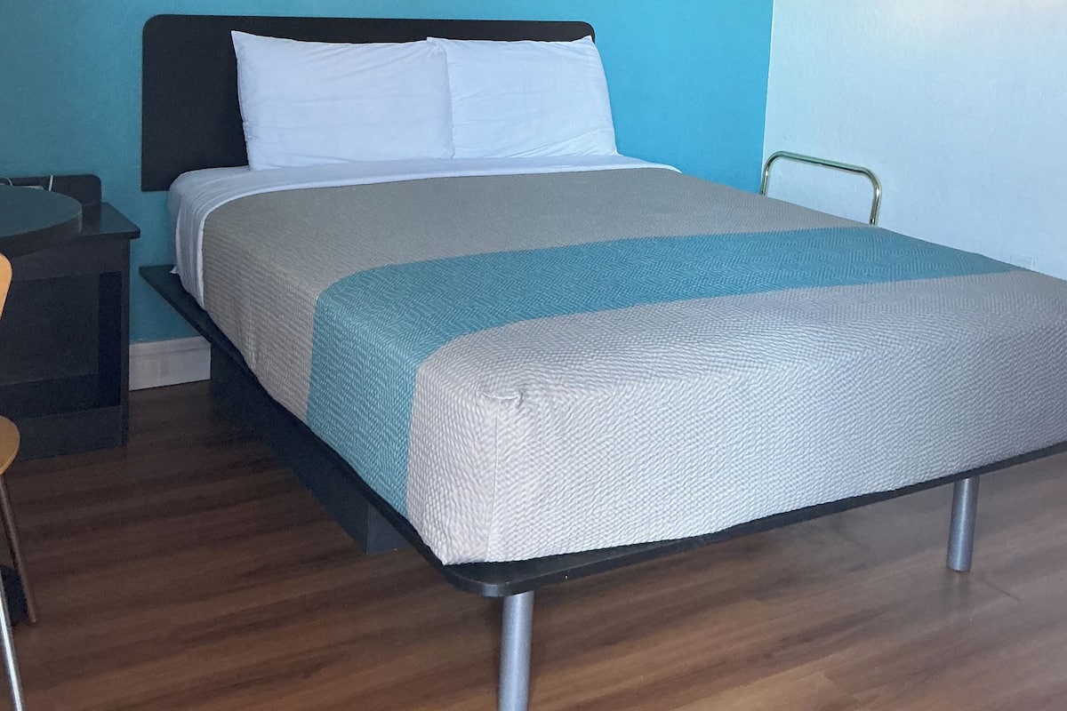 Un dormitorio con cama, mesita de noche, silla, ventilador de techo y una pared con interruptor de luz.