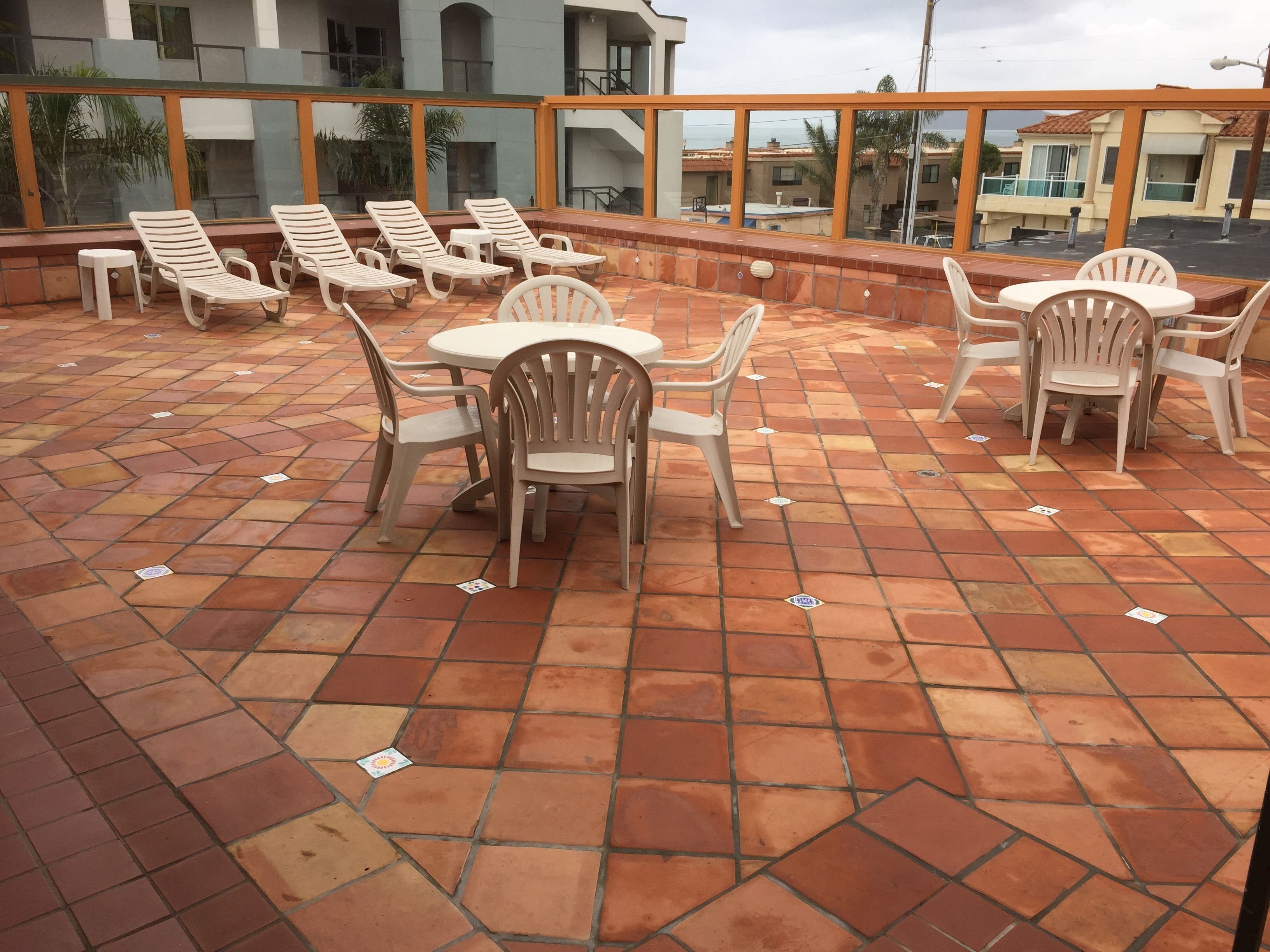 Um pátio externo com piso ladrilhado, cadeiras de plástico brancas e mesas.