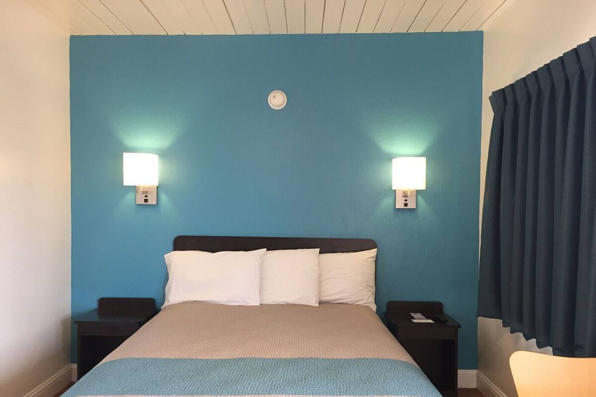 Habitación de hotel con una cama grande, dos mesitas de noche, una lámpara auxiliar y una silla, con dos luces fijadas a la pared.