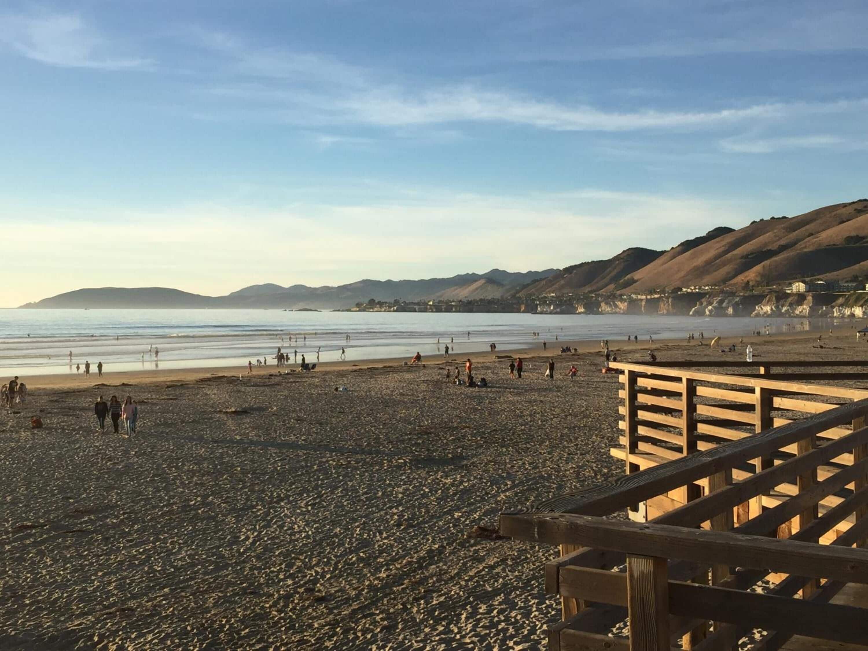 A beach with a wooden boardwalk leading to the shore, rocky terrain, and distant hills.