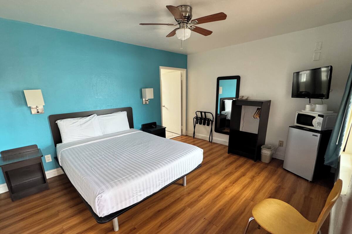 Habitación de hotel con cama, escritorio, silla, ventilador de techo, microondas y televisor.