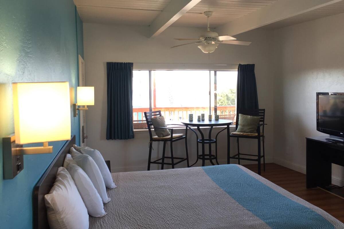 Habitación de hotel con cama, escritorio con televisor, ventilador de techo, ventana con cortinas y vista al exterior.