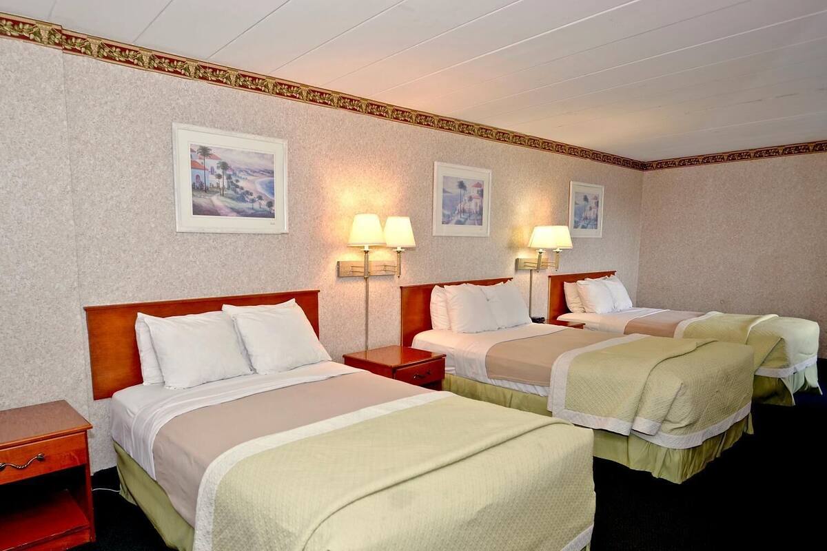 Ein Hotelzimmer mit drei Betten, jedes mit einem Holz-Kopfteil und weißer Bettwäsche. An der Wand sind Wandlampen und gerahmte Bilder angebracht.