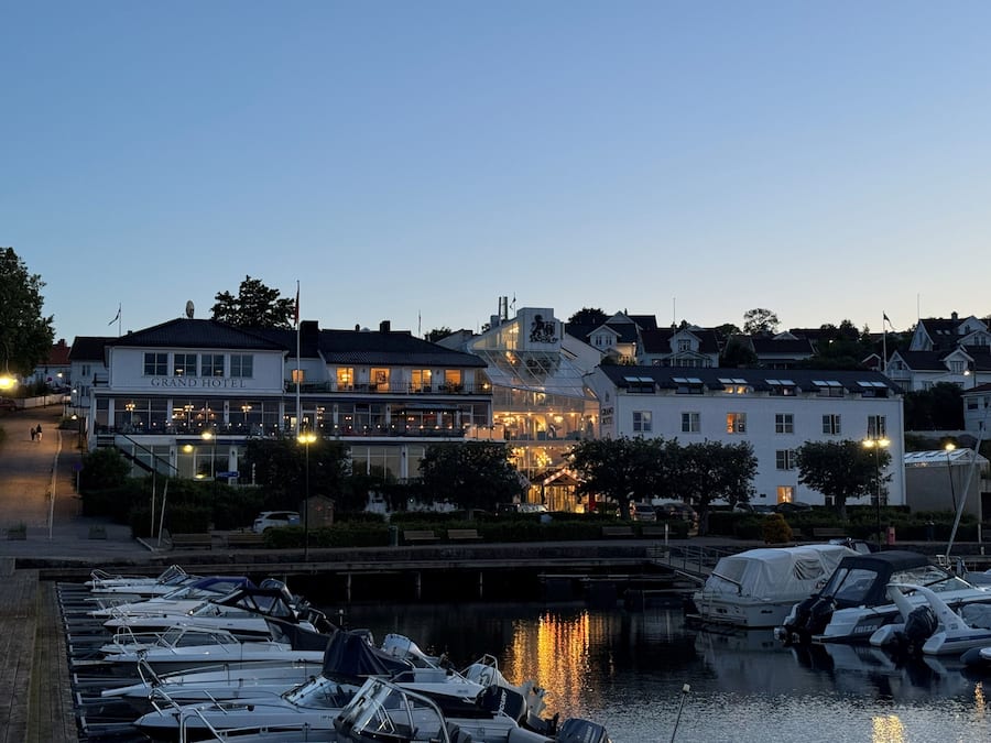A marina with boats docked, a waterfront building with a sign reading 'Grand Hotel,' and a bridge in the background.