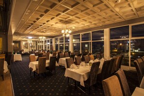Un restaurante con mesas dispuestas para comer, grandes ventanales y vista nocturna de la ciudad.