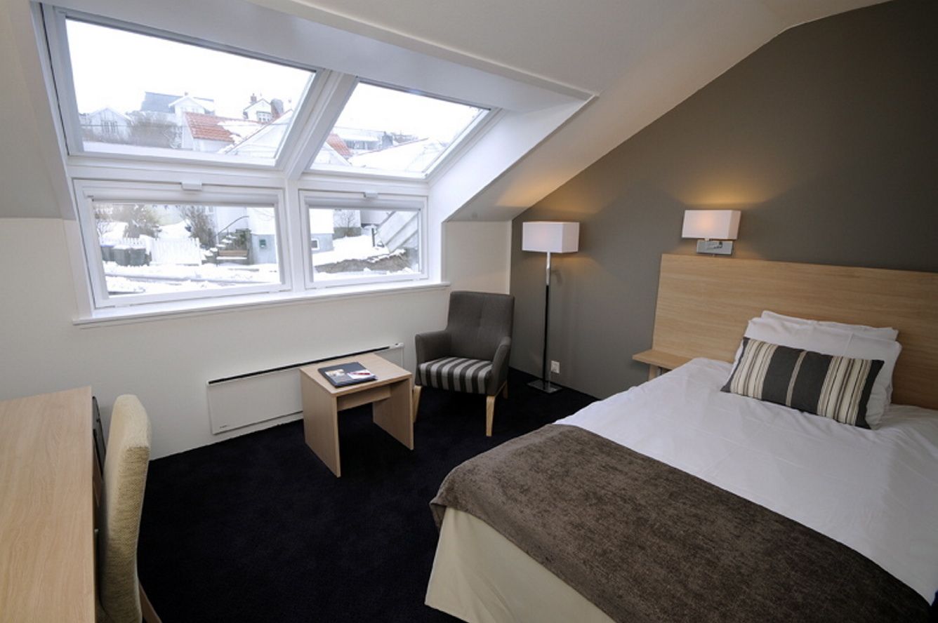 Une chambre d’hôtel avec une lucarne, un lit, un bureau, une chaise, une table de chevet et une lampe.