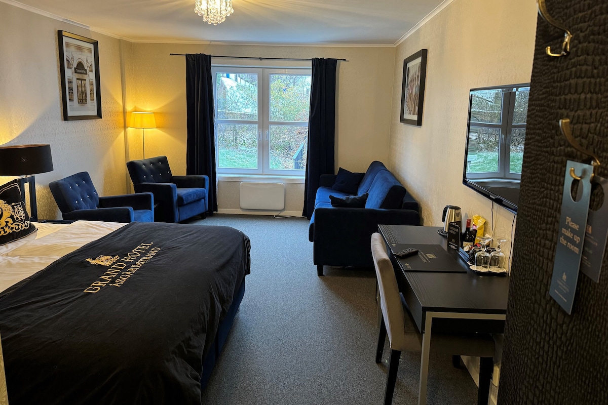 Habitación de hotel con una cama grande, dos sillones, un sofá, un escritorio con una silla, una ventana con cortinas y un candelabro.