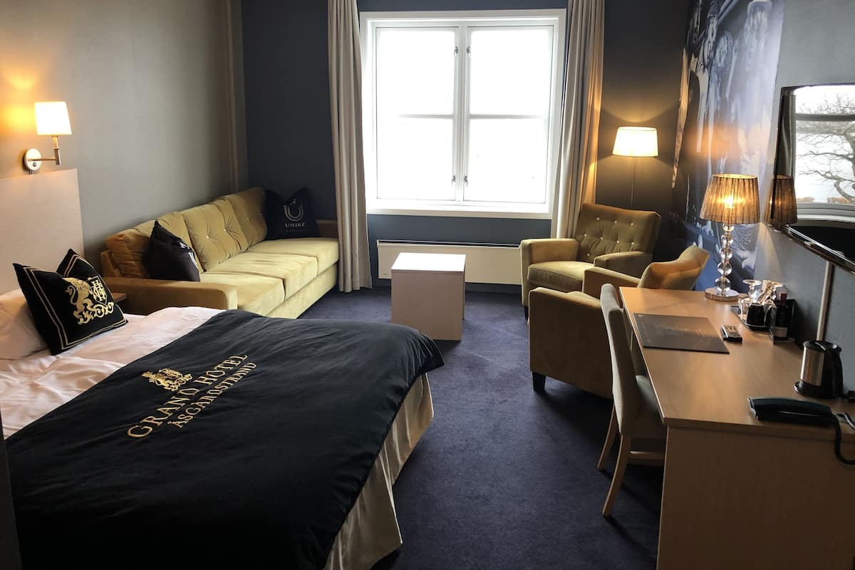 A hotel room with a bed, a sofa, a desk, and a chair. There is a window with curtains, a lamp, and a painting on the wall.