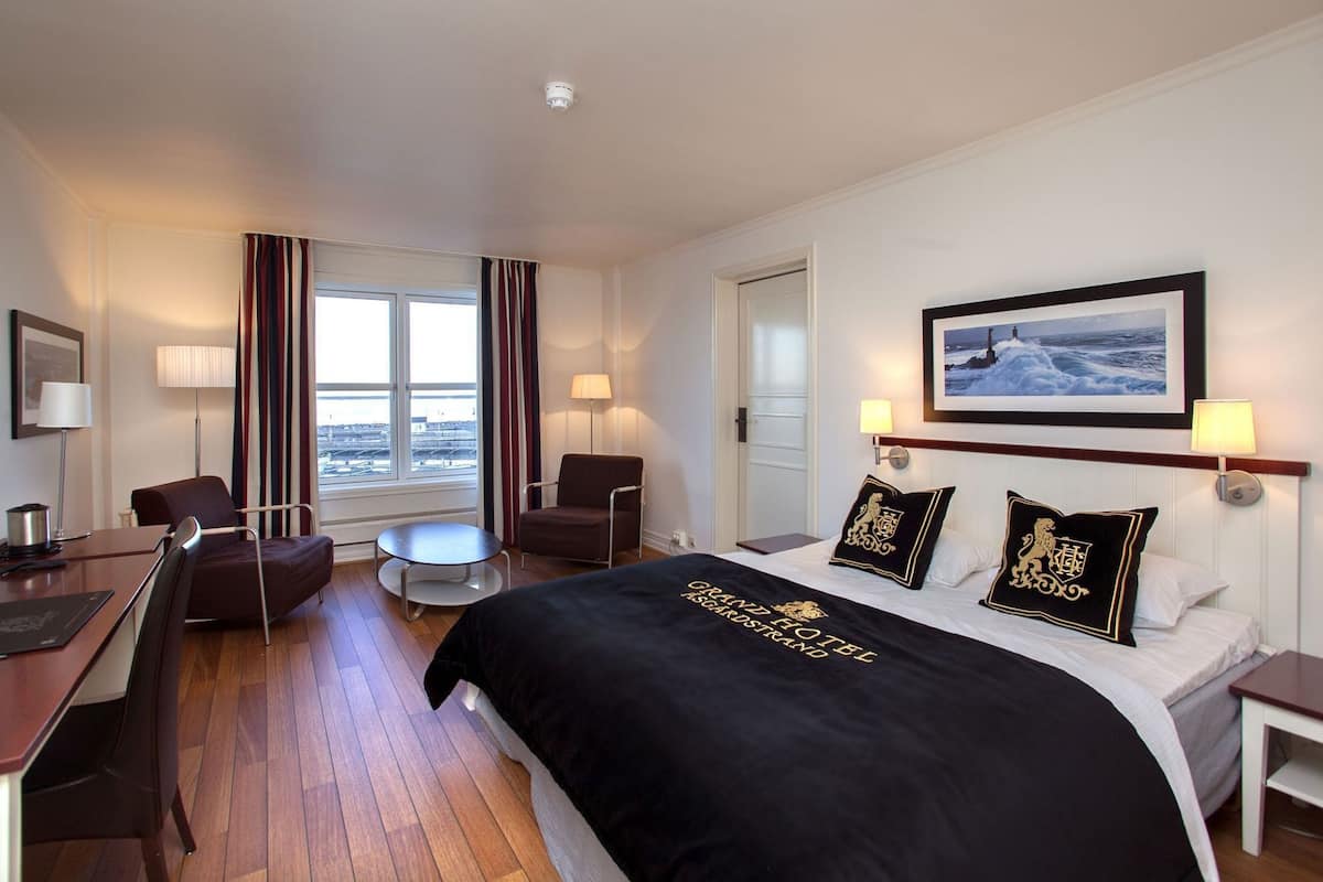 A hotel room with a bed, two chairs, a desk, a small table, and a window with curtains.