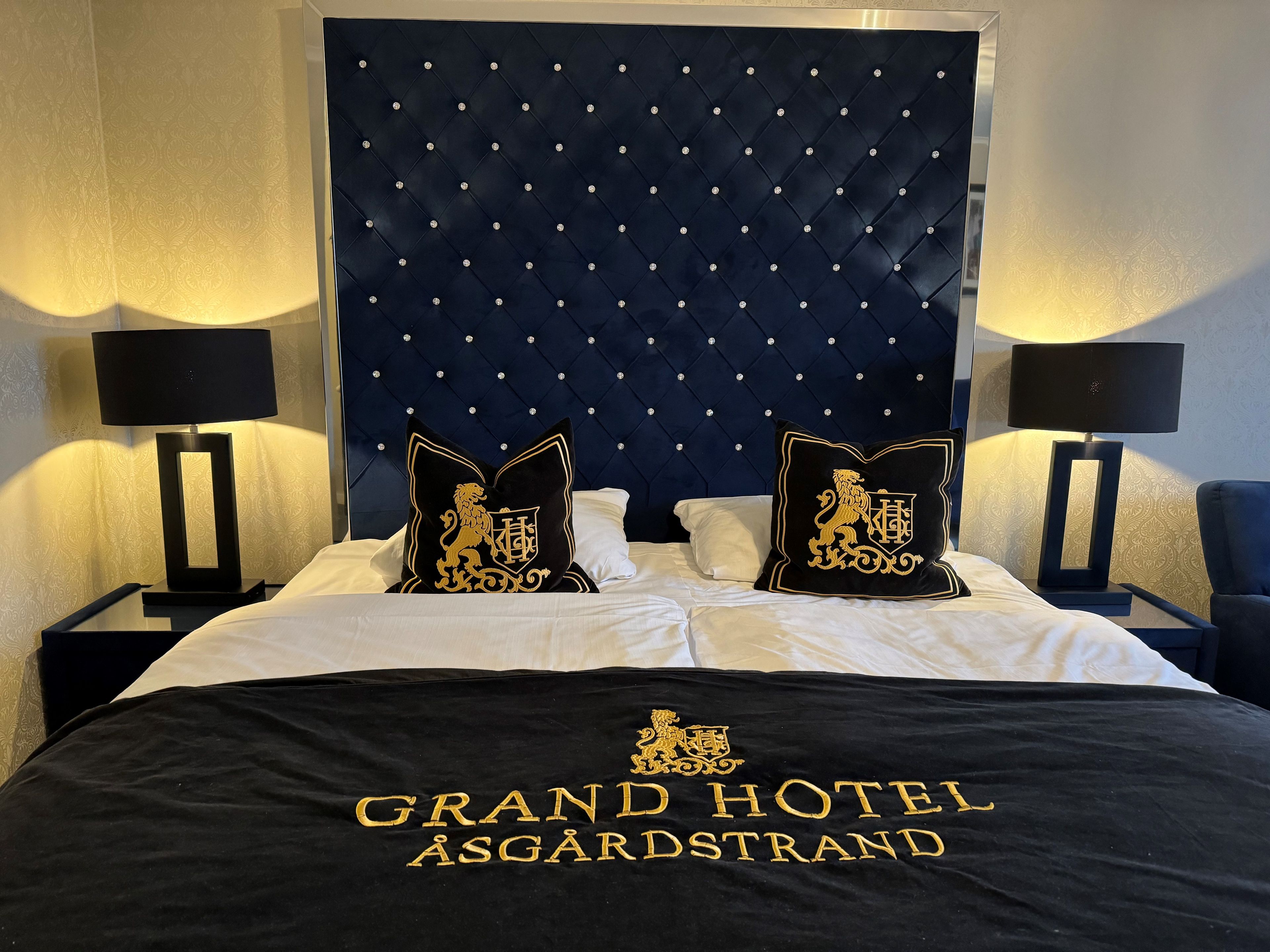 A hotel room with a tufted headboard, two black nightstands with lamps, a bed with white linens, and two decorative pillows with gold embroidery.